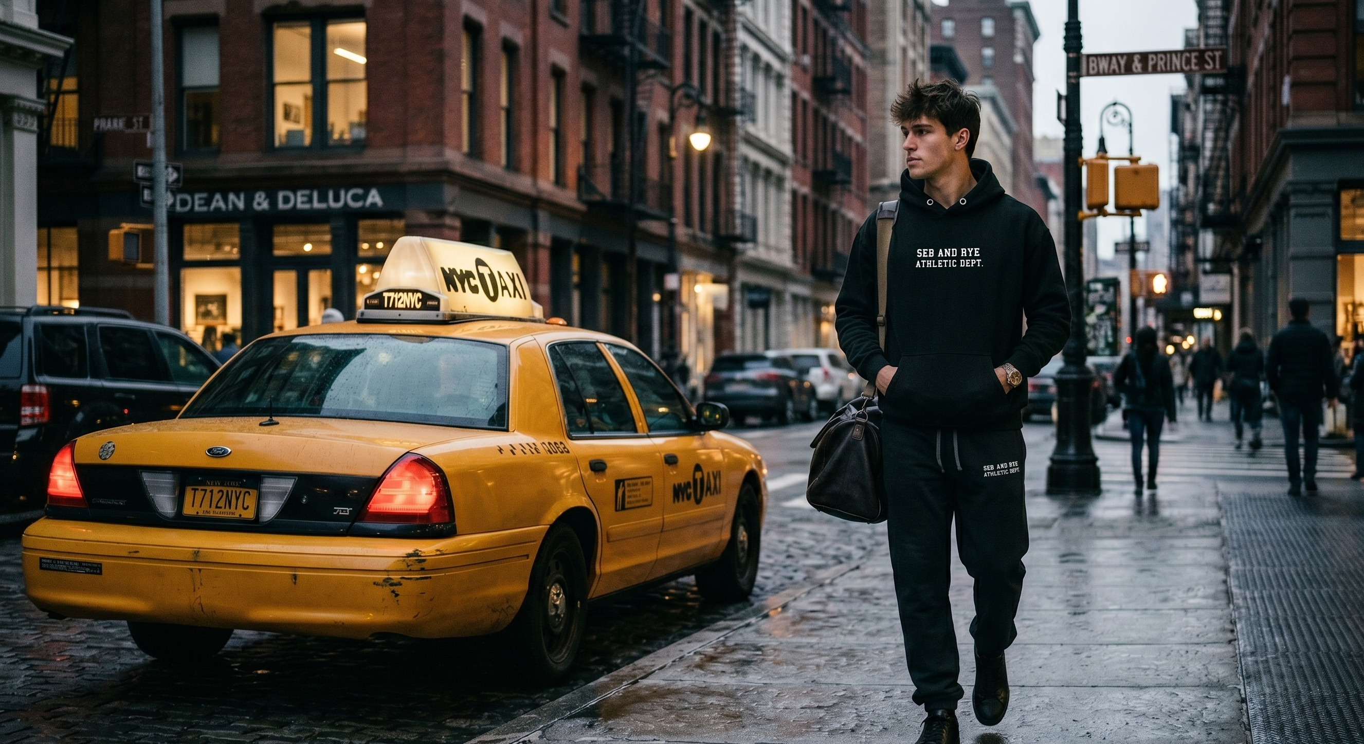 A man in a black hoodie and sweatpants walks on a city sidewalk next to a yellow taxi, with urban buildings and street details in the background. sebastian and rye