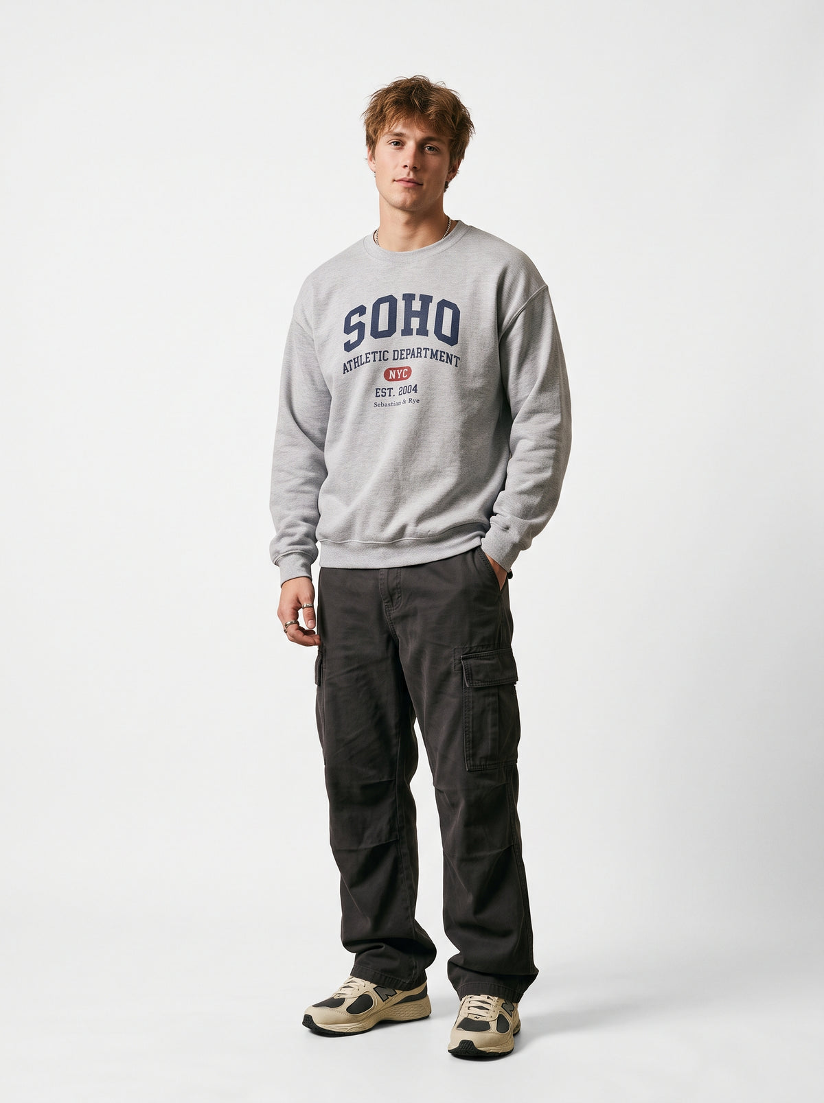 A man modeling a light gray crewneck sweatshirt with 'SOHO' printed in navy blue on the chest, paired with dark cargo pants and beige sneakers, against a plain light gray studio background.