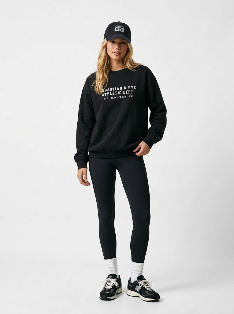 A woman modeling a black crewneck sweatshirt with white text, black leggings, a black cap, white socks, and black sneakers against a light gray studio background.