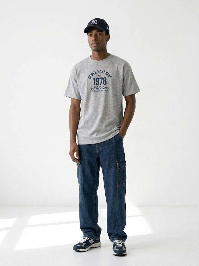 A man modeling a light gray heather crewneck t-shirt with a navy blue graphic print that reads 'SUPER EAST SIDE 1978 Fabulous', paired with dark blue jeans, a navy baseball cap, and sneakers, standing against a white studio background.